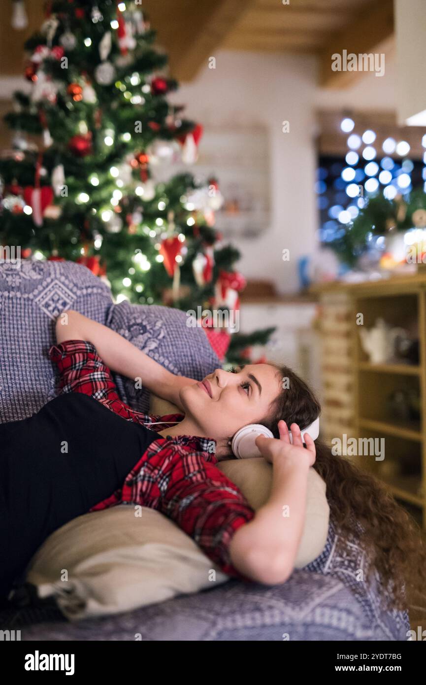 Ein Student, der Hygge Weihnachten zu Hause hat. Die Frau sitzt auf dem Sofa am Weihnachtsbaum und hört Musik mit Kopfhörern. Stockfoto