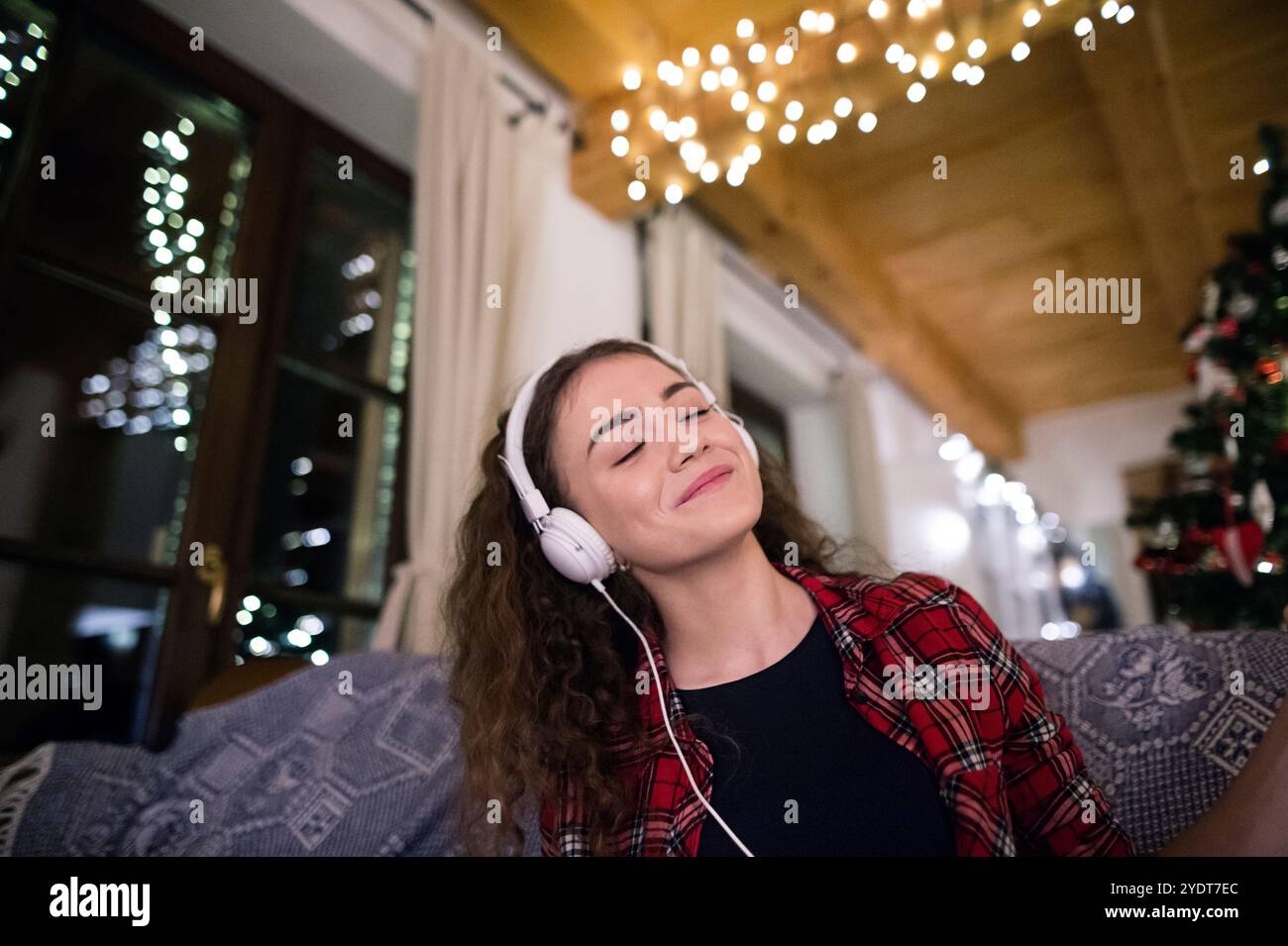 Ein Student, der Hygge Weihnachten zu Hause hat. Die Frau sitzt auf dem Sofa am Weihnachtsbaum und hört Musik mit Kopfhörern. Stockfoto