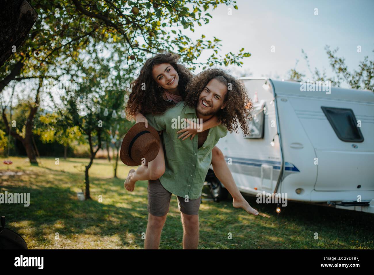 Ein verliebter Mann, der seine Verlobte mit dem Piggiback versorgt. Das junge Paar ist auf einem Campingausflug in die Natur unterwegs mit dem Wohnwagen. Stockfoto