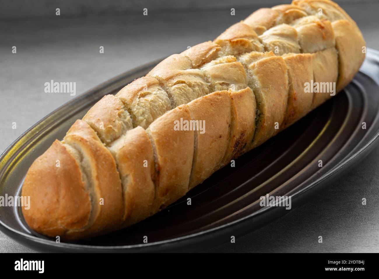 Hausgemachtes Knoblauchbrot-Baguette, vorgeschnitten auf einem schwarzen Steingut-Teller. Auf einer grauen Arbeitsfläche. Stockfoto