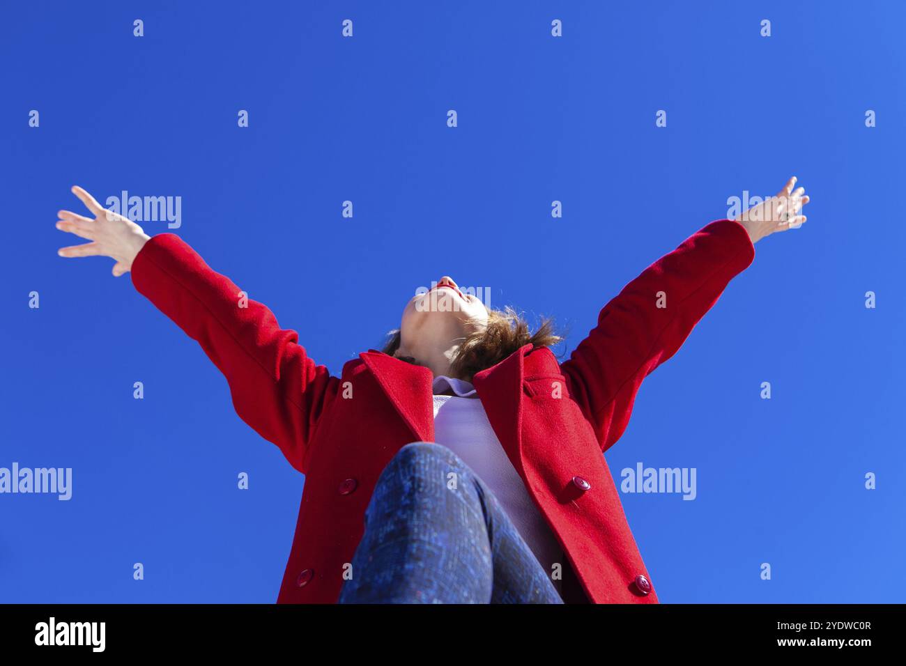 Elated kaukasische Frau, trug einen roten Mantel, hat die Arme in der Luft mit Freude an einem hellen Winter vor blauem Himmel, geschossen von einem niedrigen Winkel pro Stockfoto
