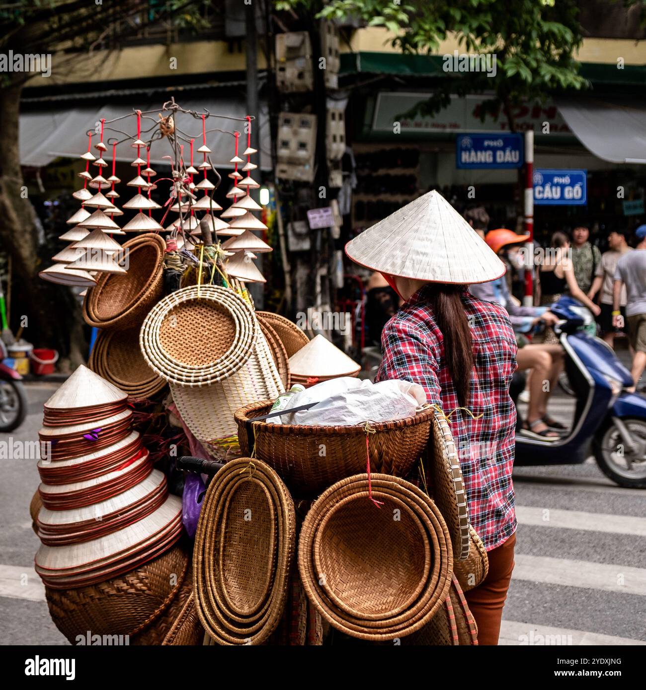 Ein Straßenverkäufer, der eine Reihe traditioneller vietnamesischer konischer Hüte und Holzkörbe verkauft, die darauf warten, eine Straße in der geschäftigen Altstadt von Hanoi zu überqueren. Stockfoto