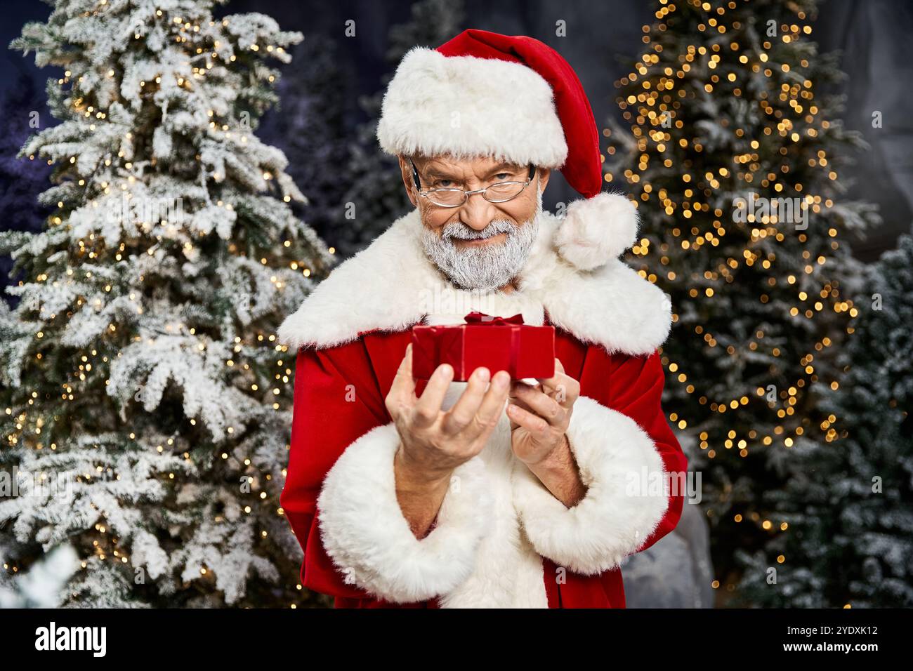 Ein fröhlicher Weihnachtsmann präsentiert ein wunderschön gewickeltes Geschenk inmitten bezaubernder schneebedeckter Bäume. Stockfoto