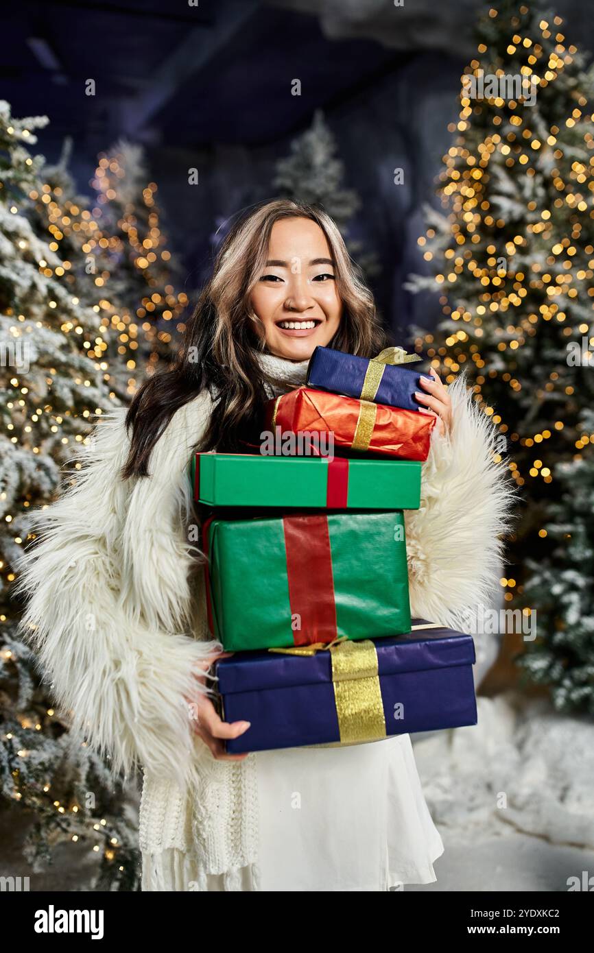 Eine junge Frau lächelt hell und hält gestapelte Geschenke in der Hand inmitten von atemberaubender Weihnachtsdekoration. Stockfoto