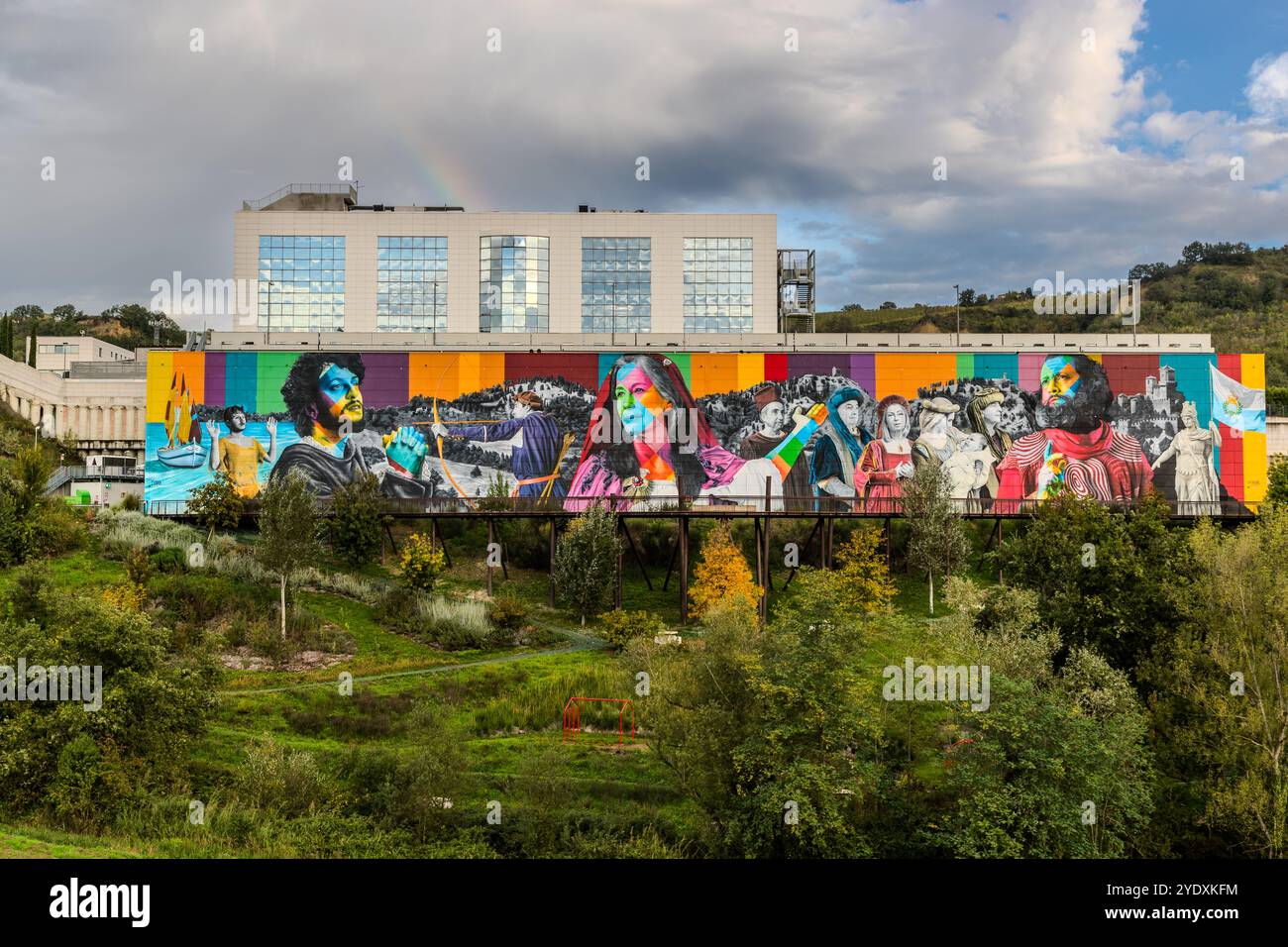 Nur ein Schotterweg führt zu diesem Blick auf das etwa 1.300 Quadratmeter große Wandgemälde "Die Geschichte von San Marino" des brasilianischen Künstlers Eduardo Kobra (2022) an der Fassade einer Verpackungsfabrik in Faetano. Im Hintergrund erheben sich die drei Türme des historischen Zentrums von San Marino, und ein Regenbogen über dem Gebäude verleiht der Szene einen magischen Moment. Strada del Sabattino, Faetano, San Marino. Stockfoto