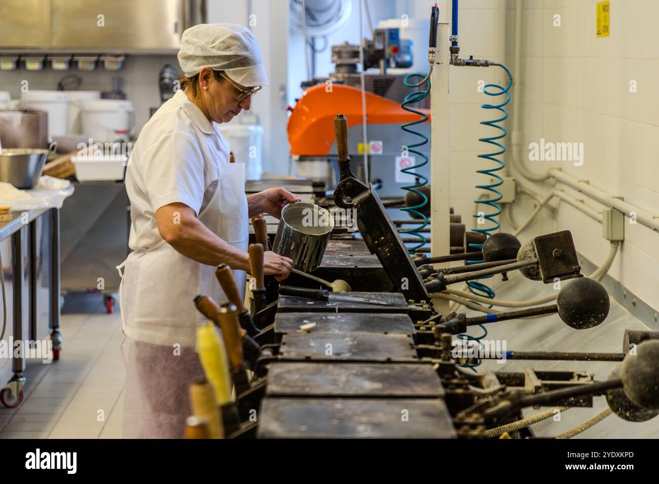 Ein Mitarbeiter gießt Waffelteig in das schwere Waffeleisen. Nach vier Minuten ist die Waffel gebacken. Die Torta Tre Monti besteht aus mehreren Lagen Waffeln und dunkler Schokolade. Ein Angestellter in weißer Arbeitskleidung und einer Kapuze füllt ein Waffeleisen in der Waffelfabrik La Serenissima. Via XXV Marzo, Domagnano, San Marino Stockfoto