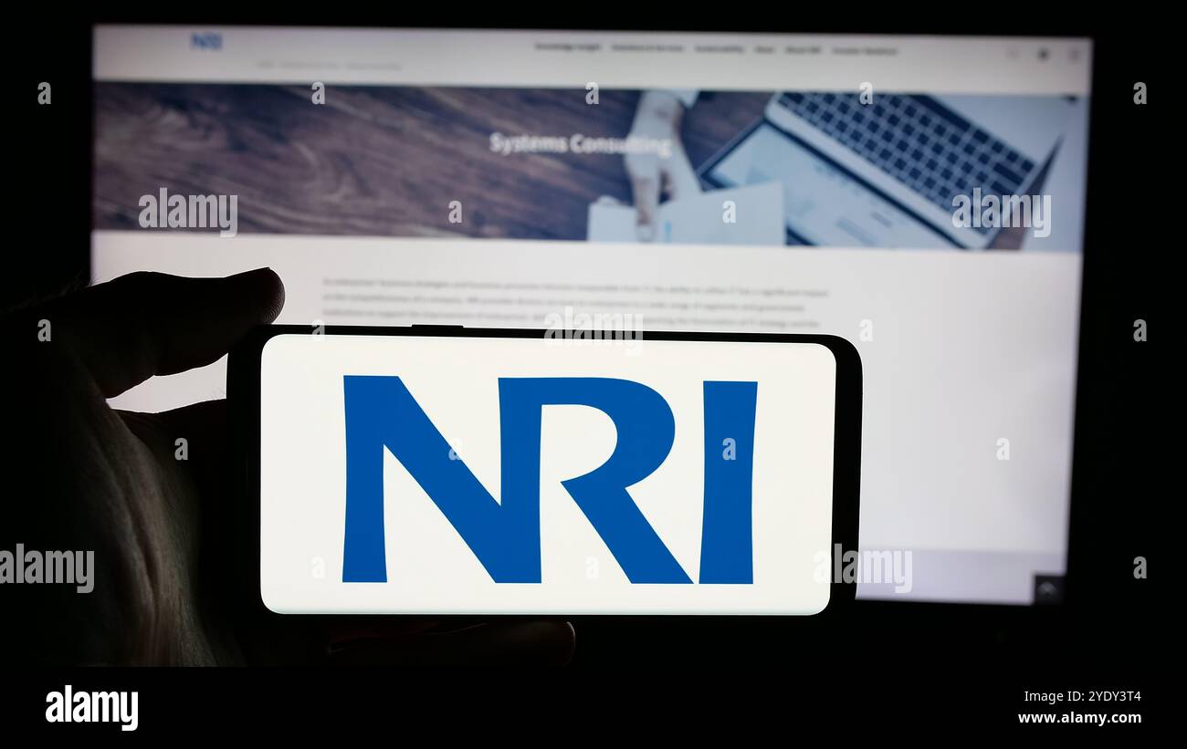 Person, die ein Mobiltelefon mit dem Logo der Firma Nomura Research Institute Ltd. (NRI) auf dem Bildschirm vor der Webseite hält. Schwerpunkt auf dem Display des Telefons. Stockfoto