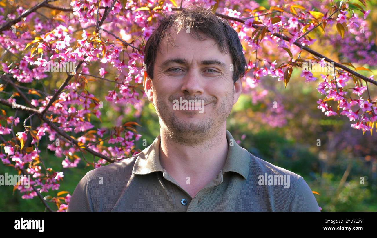 Mann lächelt unter blühenden Kirschblüten in üppigem, lebhaftem Garten, Stockfoto