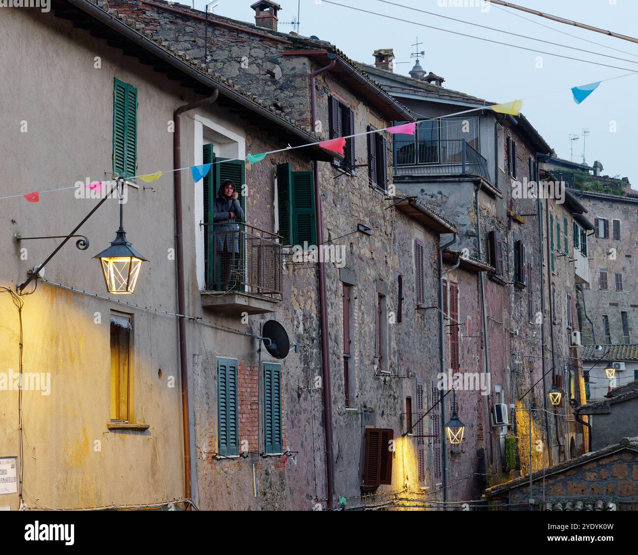Bunting und Straßenlaterne in der Abenddämmerung, während eine Dame auf einem Balkon steht, der in die historischen Häuser der Stadt Latera eingewickelt ist. Oktober 2024 Stockfoto