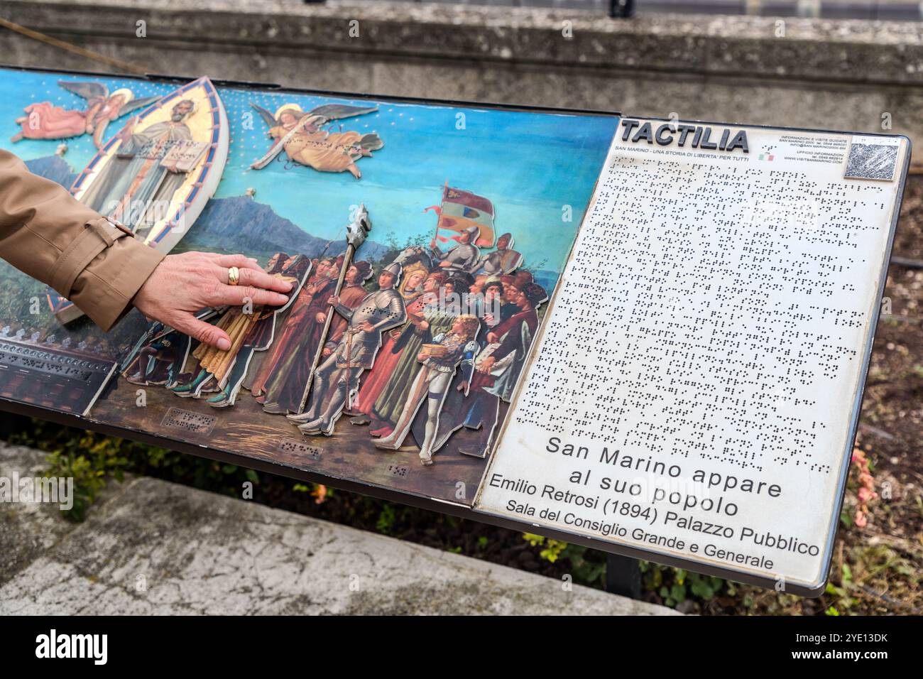 Eine Hand berührt das erhabene taktile Relief von Emilio Retrosis Gemälde von 1894 "San Marino appare al suo popolo" ("Der Heilige Marinus erscheint seinem Volk") auf einem Tactilia-Panel, dessen begleitender Text ebenfalls in Blindenschrift gedruckt ist. Das Original hängt in der Sala del Consiglio Grande e Generale des Palazzo Pubblico. Tactilia ist ein Freiluft-Taktilmuseum, das 2019 für Besucher mit Sehbehinderungen im Rahmen des Projekts San Marino Accessible Tourism eingeweiht wurde. Il Cantone, Stadt San Marino, San Marino. Stockfoto
