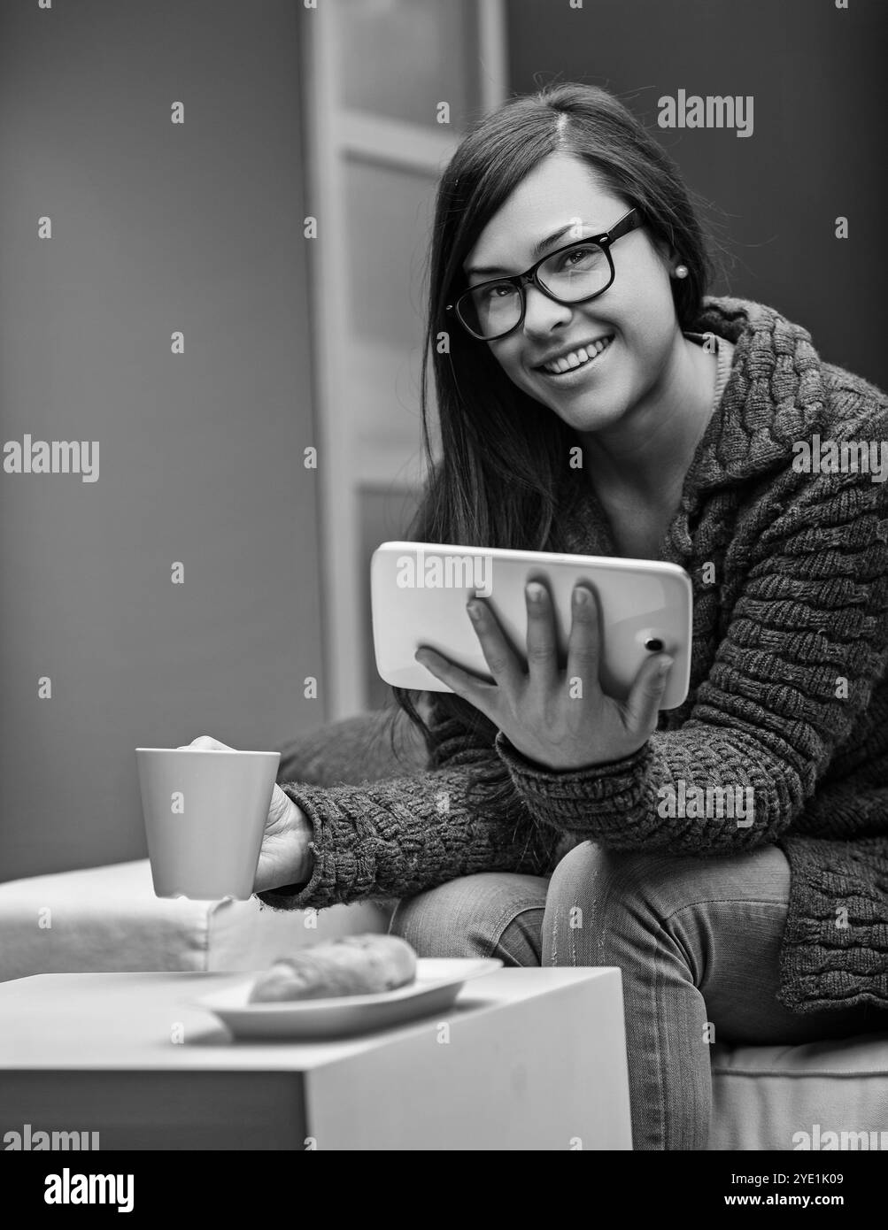 Die junge Frau entspannt sich auf dem Sofa mit einem Tablet und Kaffee und genießt einen ruhigen Moment zu Hause. Sie sieht zufrieden aus, in Technologie und warmem Bevera Stockfoto