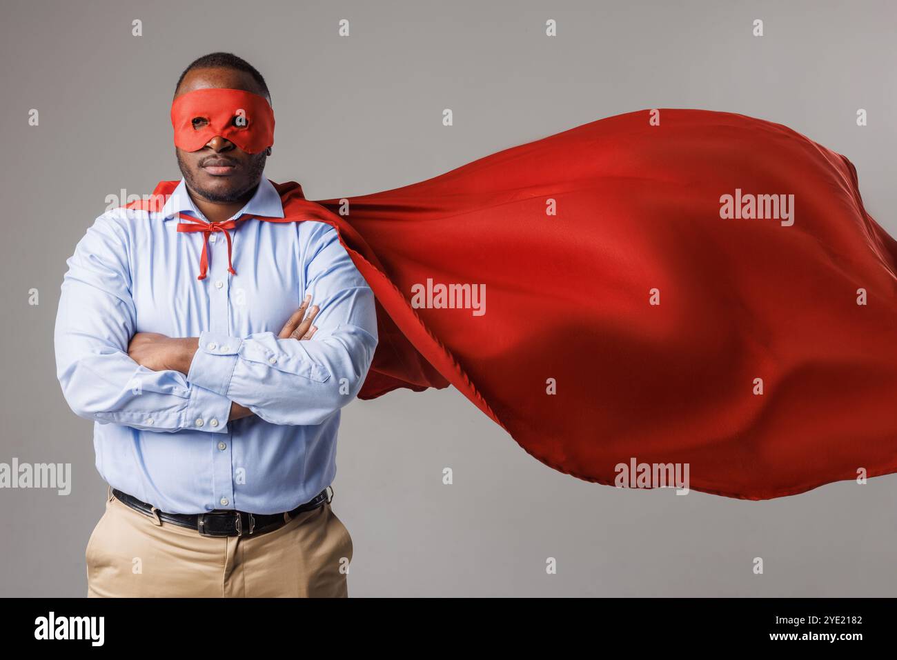 Ernster afroamerikanischer Mann im Superheldenkostüm, der seine Arme isoliert auf weißem Hintergrund faltet. Dunkelhäutiger Kerl in roter Maske und cape, super Power Co Stockfoto