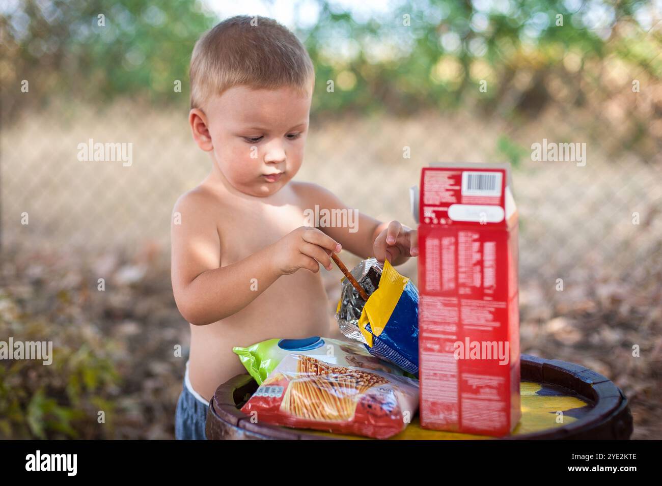 Ein kleines Kind ohne Hemd greift neugierig in einen Snackbeutel, während es in der Nähe eines Fasses sitzt. Die Umgebung bietet viel Grün und eine warme, sonnige Atmosphäre Stockfoto