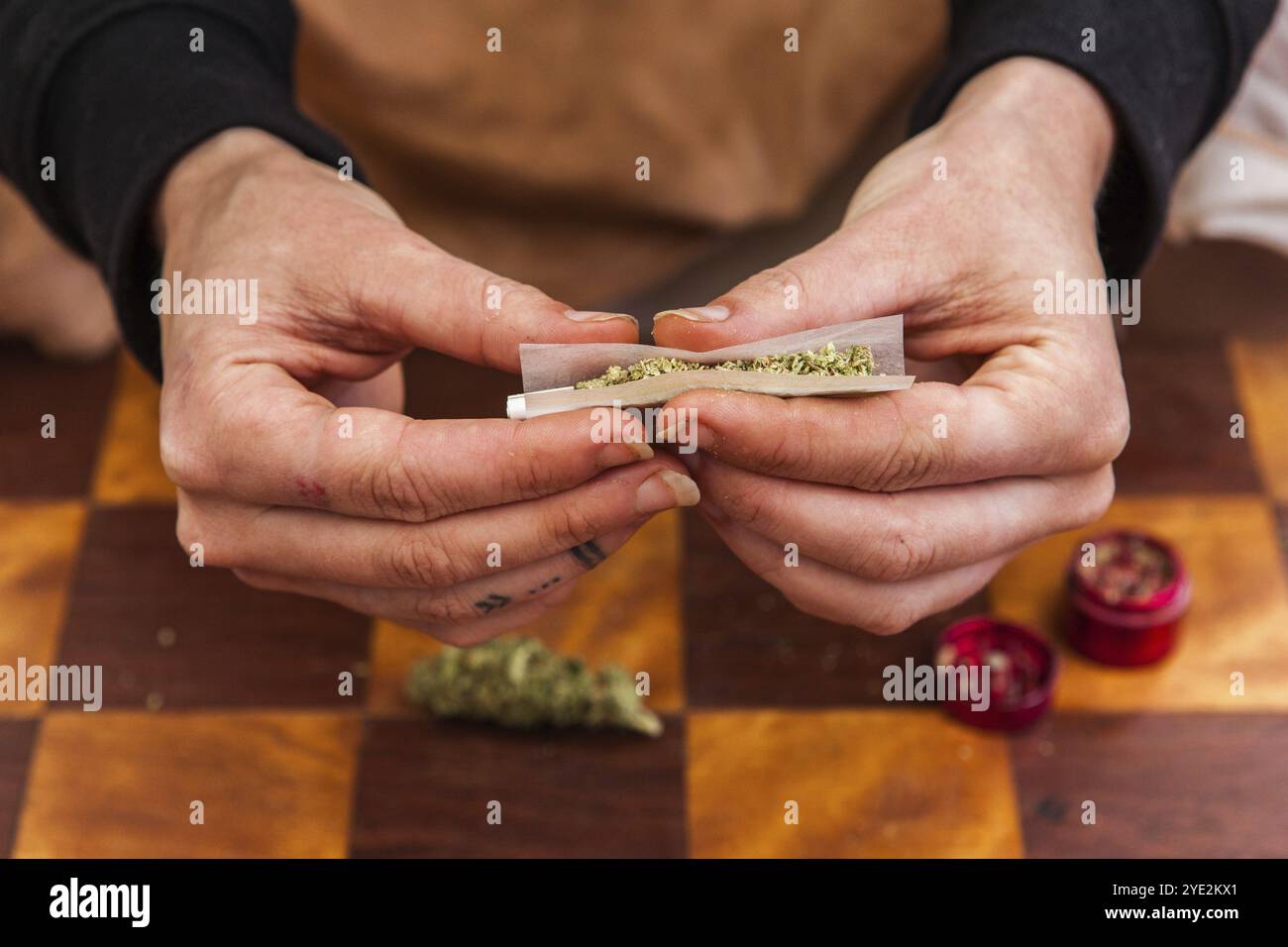 Nahaufnahme von Händen, die ein gemahlenes, mit Cannabis gefülltes Papierrollenpapier kneifen, um es in einen Joint zu Rollen. Unkrautknospen und Mahlwerk auf dem Tisch unten Stockfoto