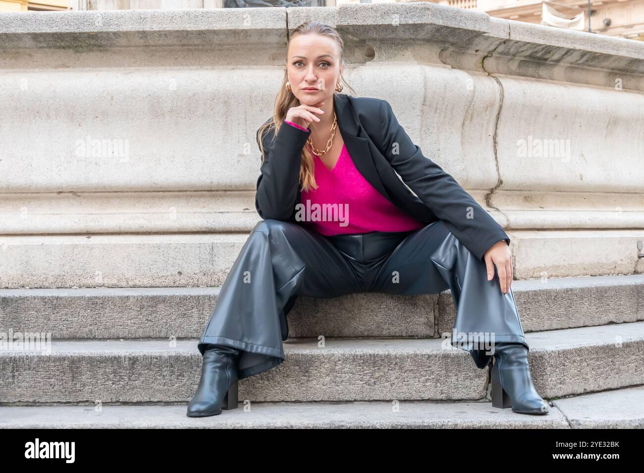 Eine selbstbewusste 35-40-jährige Frau posiert stilvoll in einem schwarzen Blazer und einem pinkfarbenen Oberteil, während sie auf Steintreppen sitzt und die urbane Mode in einem urbanen Stil betont Stockfoto