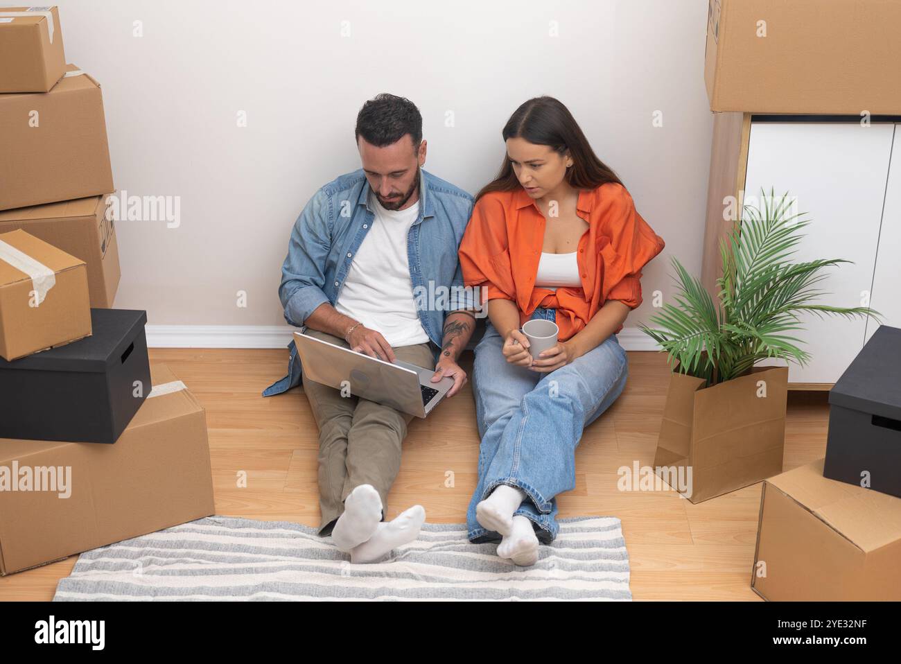 Fröhlicher junger Mann und Frau, die sich auf dem Boden ihres neuen Hauses mit Laptop entspannen. Kartons ausgepackt Stockfoto