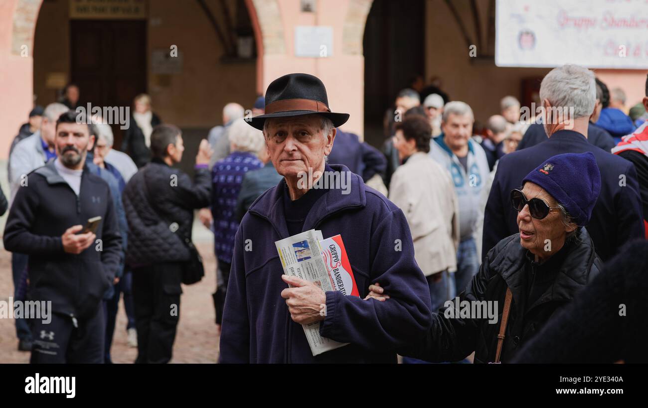 Ein älterer Mann mit Hut hält eine Zeitung und steht unter einer Menge in Alba, Italien. Der lebhafte Markttag zeigt Einheimische, die die Atmosphäre und ich genießen Stockfoto