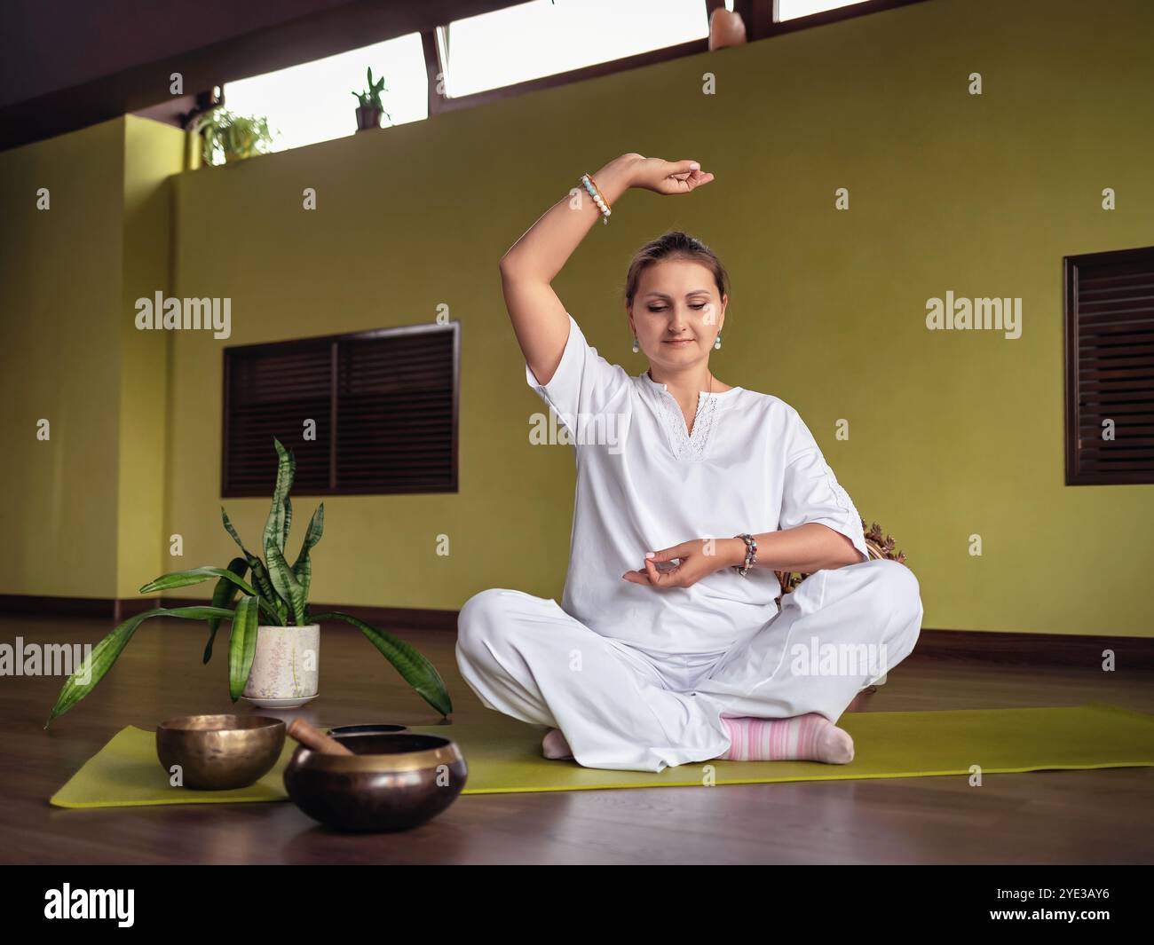 Eine Frau übt Meditation im Kundalini Yoga aus, sitzt in einer bequemen Lotusposition auf einer Matte in einem Raum, in weißer Kleidung, mit den Händen, die das zeigen Stockfoto