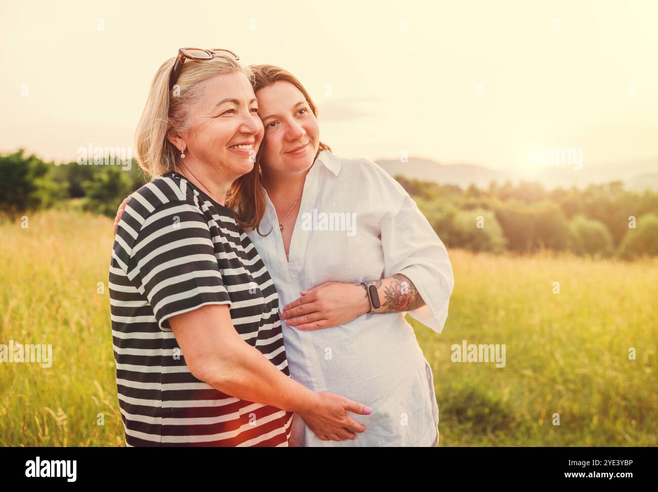 Porträt einer aufrichtig lächelnden, jungen schwangeren Frau in hellen Sommerkleidung, die abends bei Sonnenuntergang mit der Mutter umarmt. Die Gesundheit der Frau, hu Stockfoto