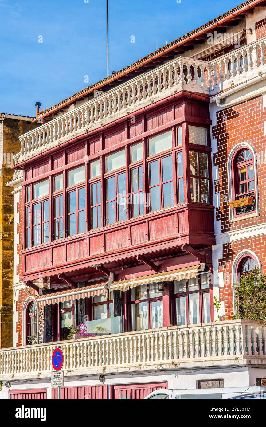 Ein rot gestrichener Holzbalkon erstreckt sich von der oberen Etage eines traditionellen Gebäudes in spanischen Fischereivierteln. Es hat viele Fenster, einen gestreiften Markisen Stockfoto