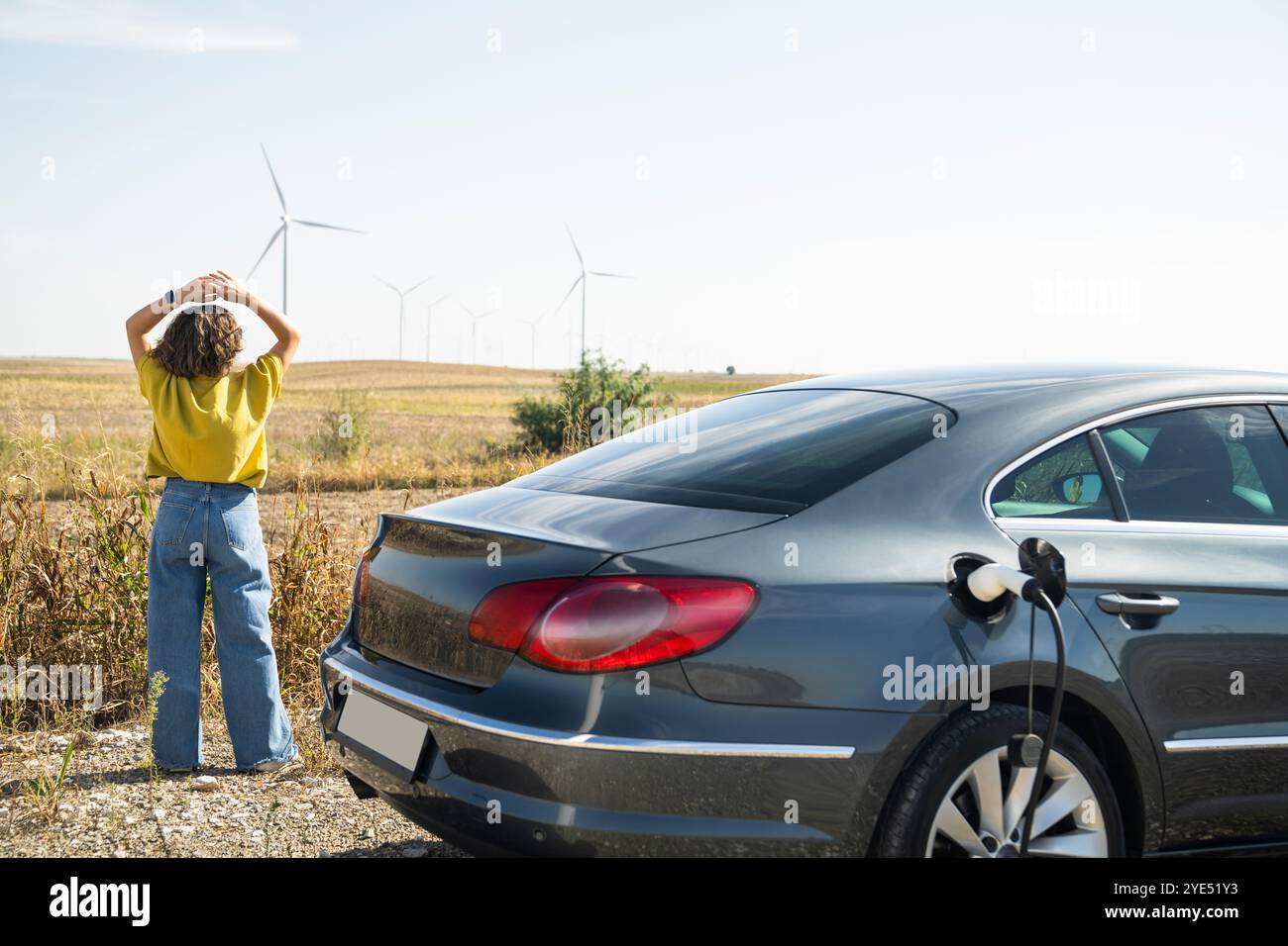 Frau mit gebogenen Haaren, die ein gelbes Hemd trägt, steht neben dem Aufladen des Elektroautos. Windräder im Hintergrund. Stockfoto