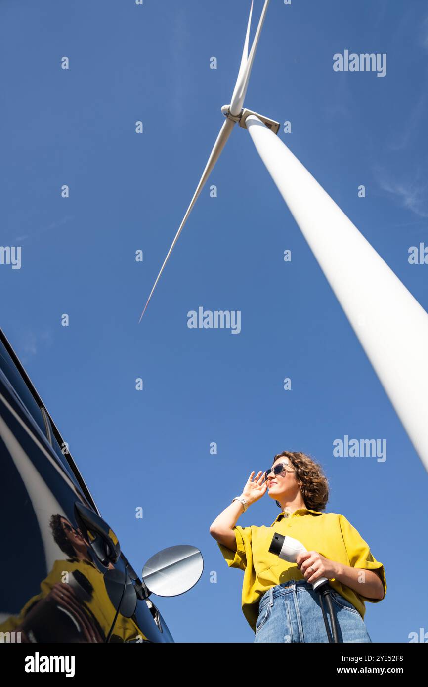 Eine Frau mit gewölbten Haaren, die ein gelbes Hemd und eine Sonnenbrille trägt, steht neben dem Aufladen des Elektroautos. Windkraftanlage im Hintergrund. Stockfoto