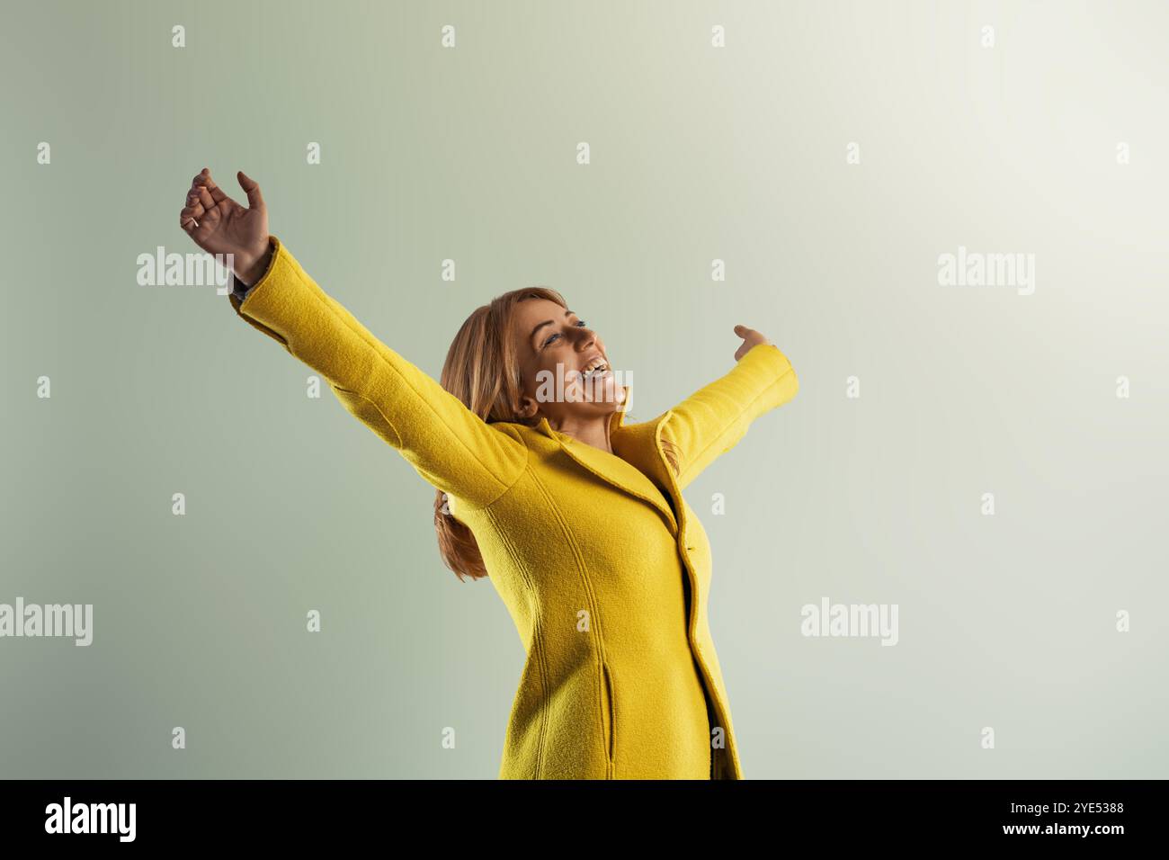 Eine fröhliche Frau in einem hellgelben Mantel hebt die Arme. Strahlende Freiheit und Glück. Freude und Positivität ausstrahlen. Hoffnung und Inspiration verkörpern. C Stockfoto