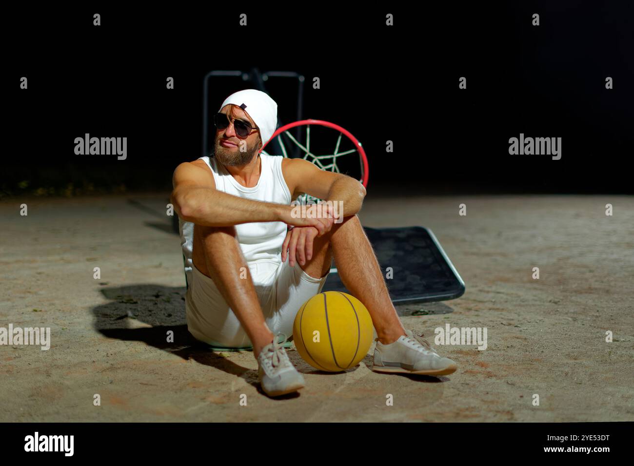 Ein Basketballspieler in weißem Outfit sitzt mit einem Ball in der Hand auf dem Platz, trägt eine Sonnenbrille und sieht nach einem harten Training entspannt aus. Stockfoto