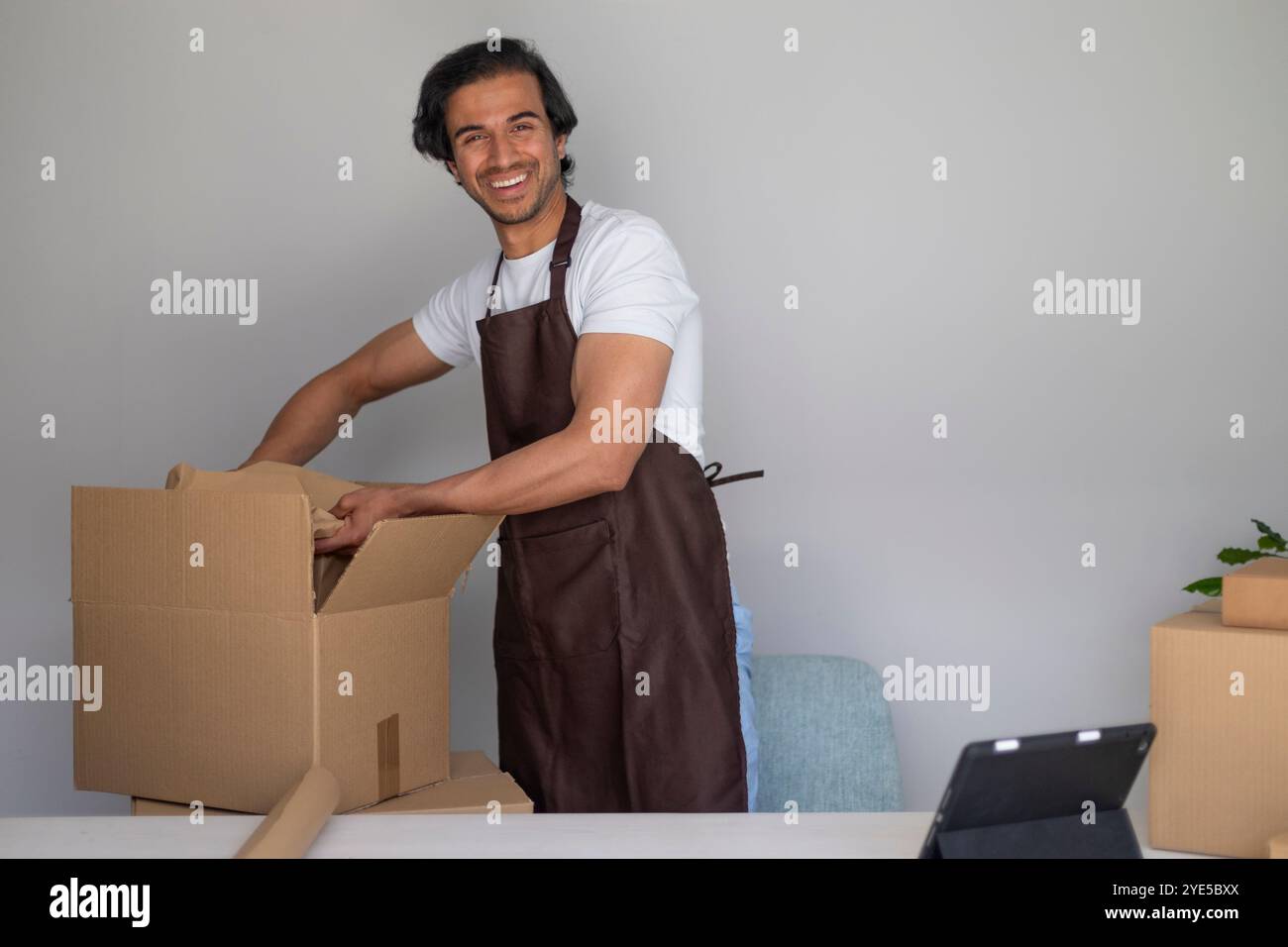 Ein Mann auf der Schürze packt glücklich einen Karton an einem Tisch, umgeben von Verpackungsmaterial, das in die Kamera lächelt Stockfoto