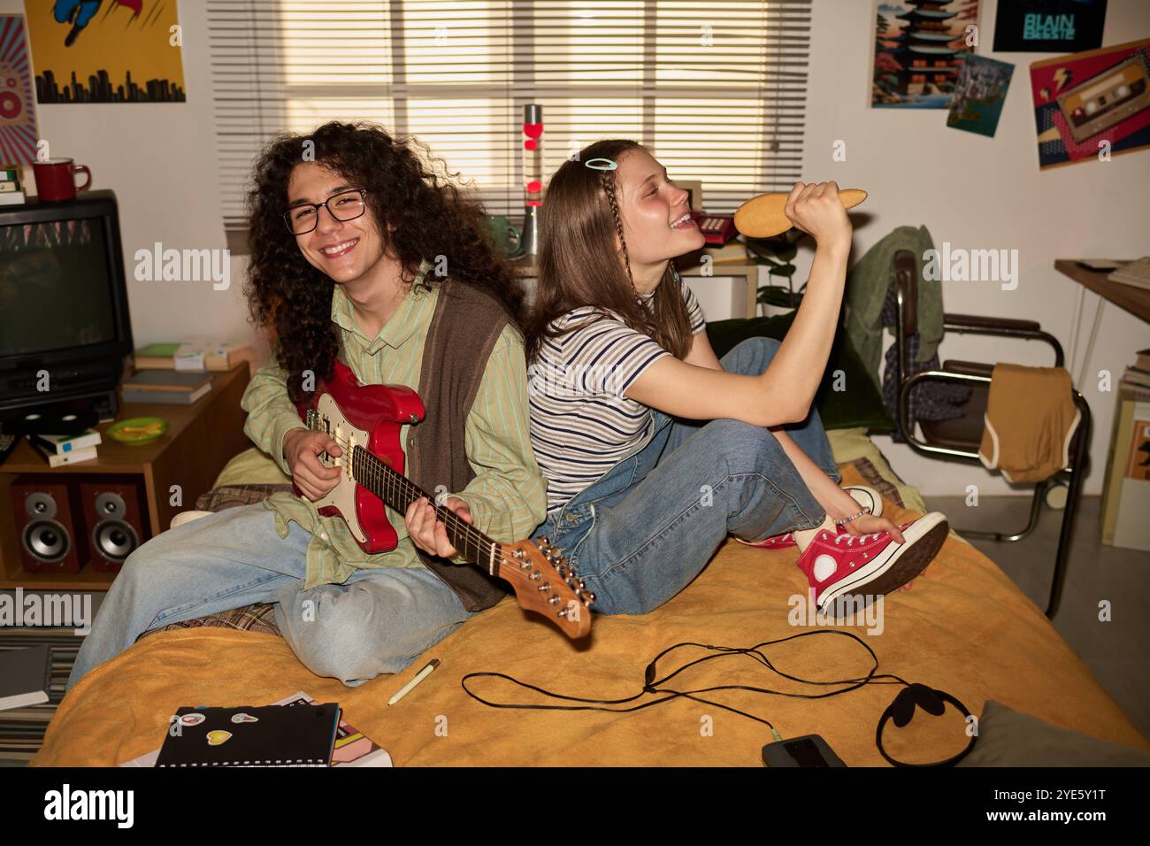 Zwei Freunde spielen Gitarre und singen fröhlich, während sie auf dem Bett sitzen, in einem gemütlichen Retro-Schlafzimmer mit Musikplakaten und Ausrüstung verstreut Stockfoto Zwei Freunde spielen Gitarre und singen fröhlich, während sie auf dem Bett sitzen, in einem gemütlichen Retro-Schlafzimmer mit Musikplakaten und Ausrüstung verstreut Stockfoto