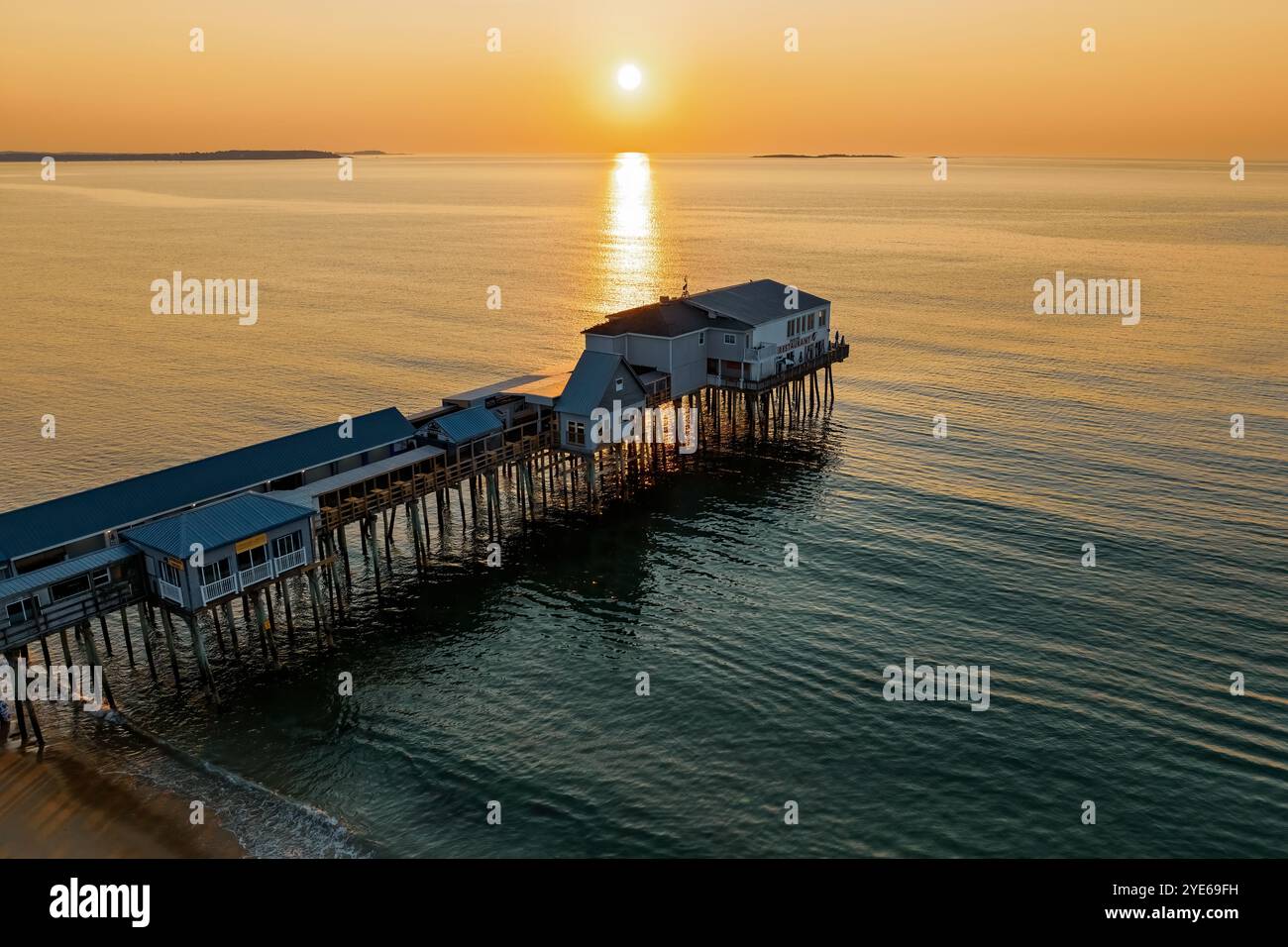 Der Old Orchard Beach Pier erstreckt sich unter einem warmen Sonnenaufgang in ruhiges Wasser, mit Gebäuden über dem Meer auf Stelzen. Das Sonnenlicht reflektiert das o Stockfoto