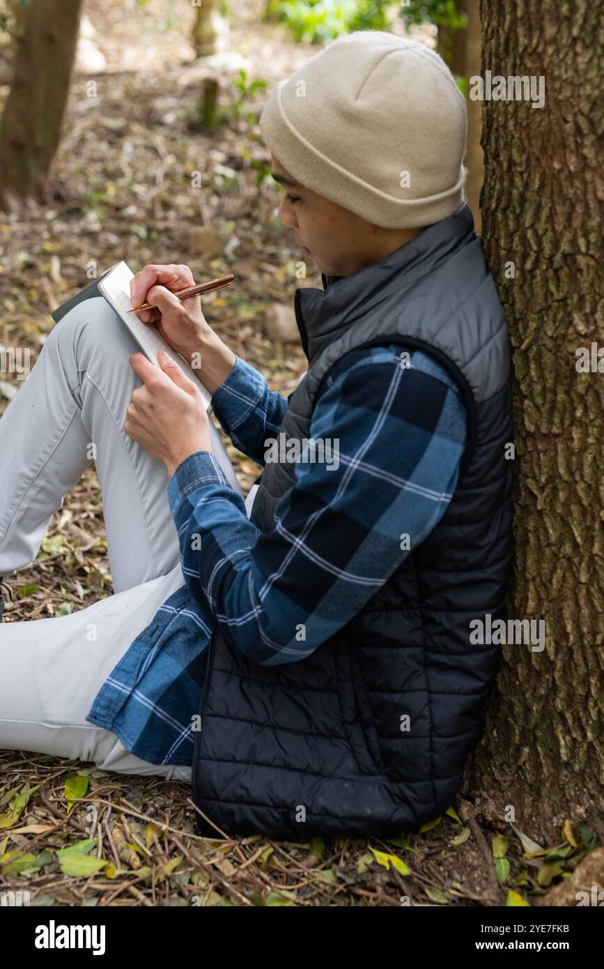 Mann, der draußen im Notizbuch schreibt, sich im Wald an einen Baum lehnt Stockfoto