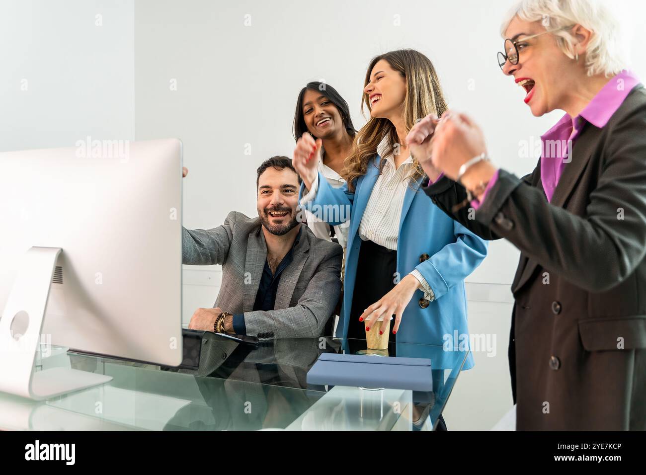 Begeisterte Geschäftsleute, die ihren Erfolg feiern, während sie Computermonitore in modernen Büros betrachten. Junge und ältere Mitarbeiter teilen Freude Stockfoto
