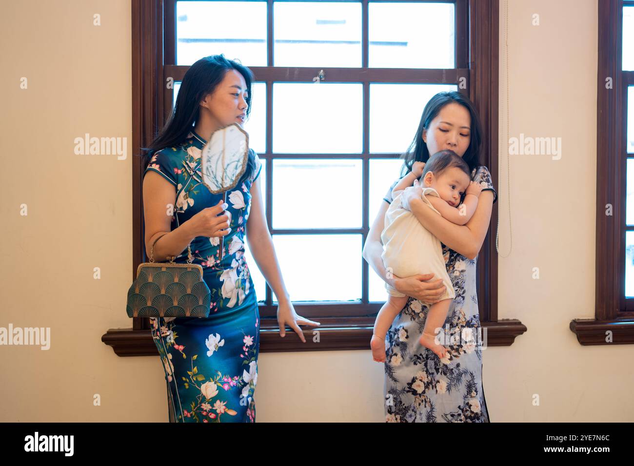 Zwei taiwanesische Frauen im Alter von 30 Jahren in traditioneller chinesischer Kleidung halten ein 6 Monate altes Baby und unterhalten sich am Tisch am Fenster des traditionellen Gebäudes Stockfoto