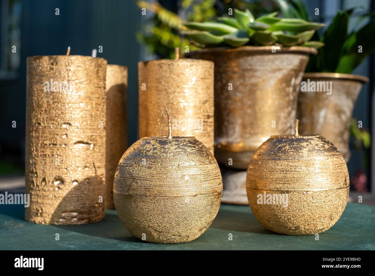 Luxuriöse goldene Kerzen in verschiedenen Formen stehen neben dekorativen Pflanzgefäßen, die alle eine stilvolle, in warmes Sonnenlicht getauchte Umgebung im Freien hervorheben. Stockfoto
