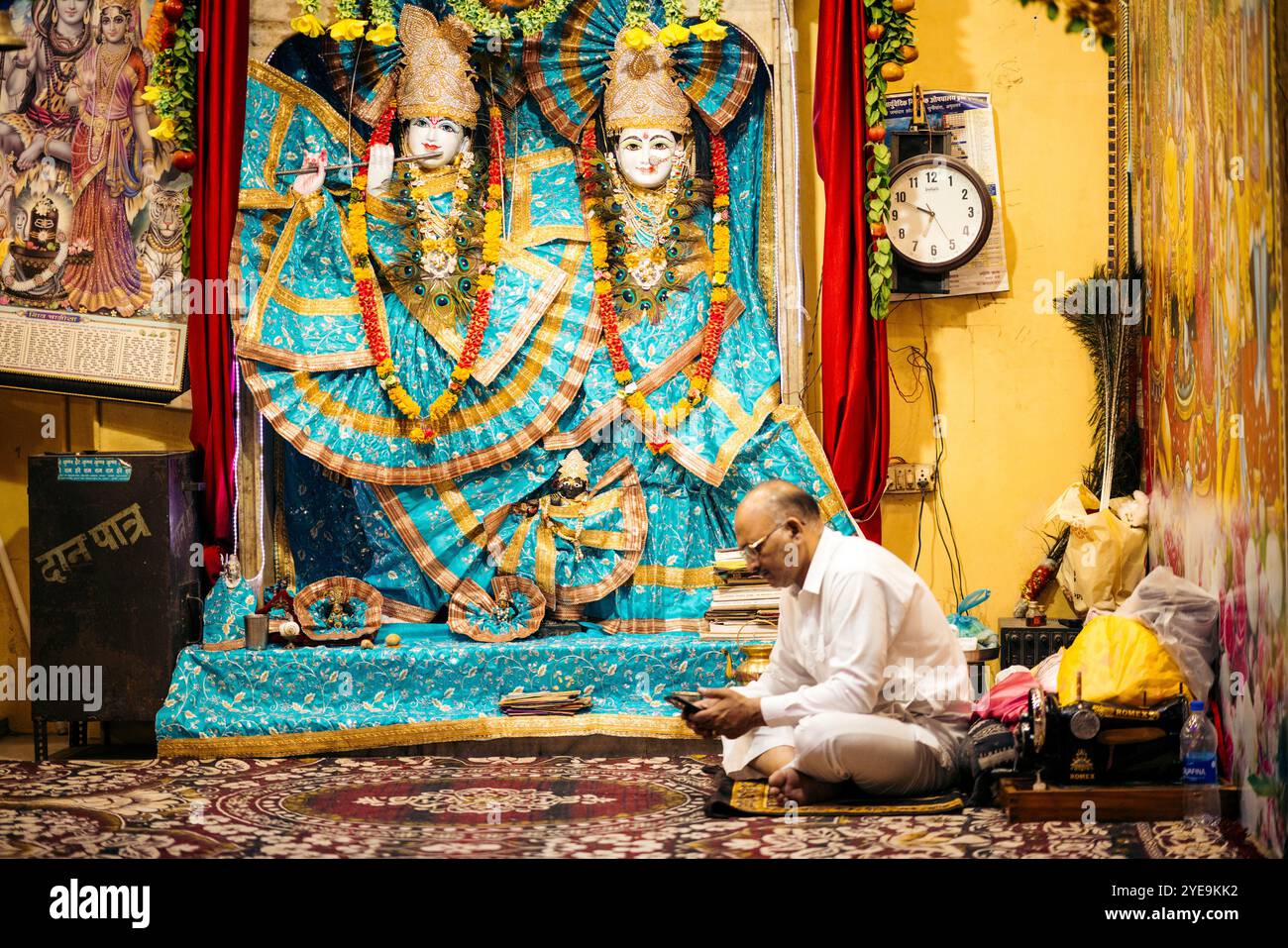 Der Mann sitzt mit dem Kreuz auf einer Matte und benutzt sein Smartphone neben einem religiösen Schrein; Amritsar, Punjab, Indien Stockfoto