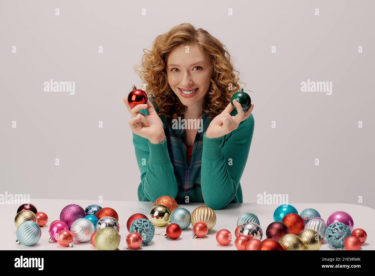 Eine glückliche junge Frau hält zwei Ornamente, während sie von festlichen Dekorationen auf einem Tisch umgeben ist. Stockfoto