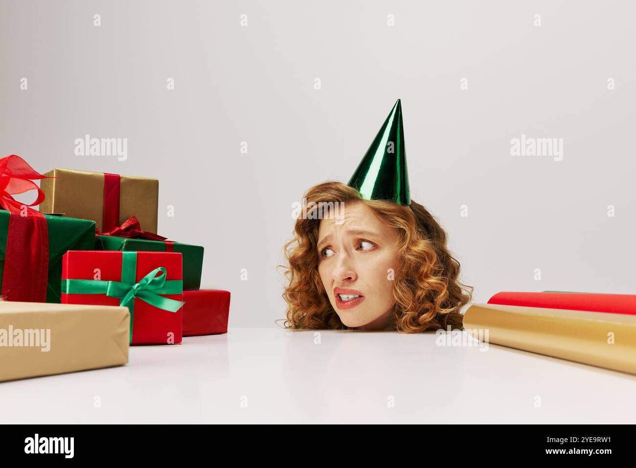 Eine junge Frau mit grünem Partyhut schaut neugierig auf bunte, gewickelte Geschenke auf dem Tisch. Stockfoto