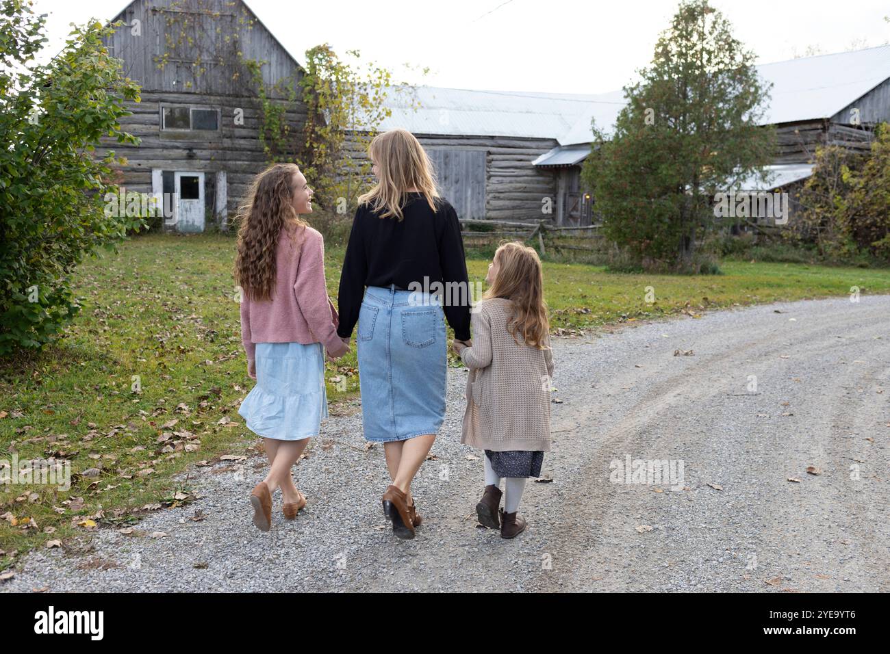Blick von hinten auf eine Mutter, die mit ihren beiden Töchtern die Hände hält, während sie auf einem Schotterweg zu einem alten hölzernen Bauernhaus in einem ländlichen... Stockfoto