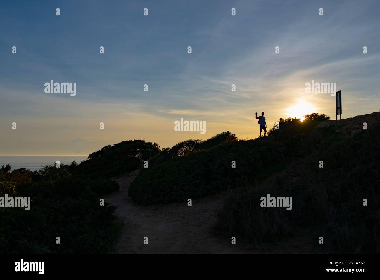 Die Person steht und blockiert die Sonne vor seinen Augen, während er über das Meer und den Sonnenuntergang an einem hohen Punkt über dem Benagil Beach entlang der Küste Portugals blickt Stockfoto