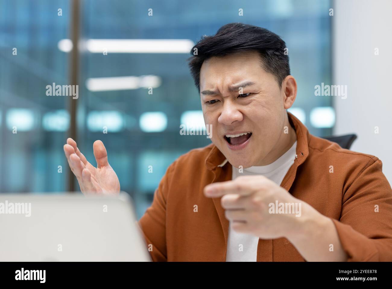 Asiatischer Mann, der Wut während eines Videoanrufs im Büro zeigt. Geschäftsleute, die ein Notebook verwenden, kommunizieren Frustration, Zeigen und Gesten. Konzept von Stress am Arbeitsplatz und Remote Meetings. Stockfoto