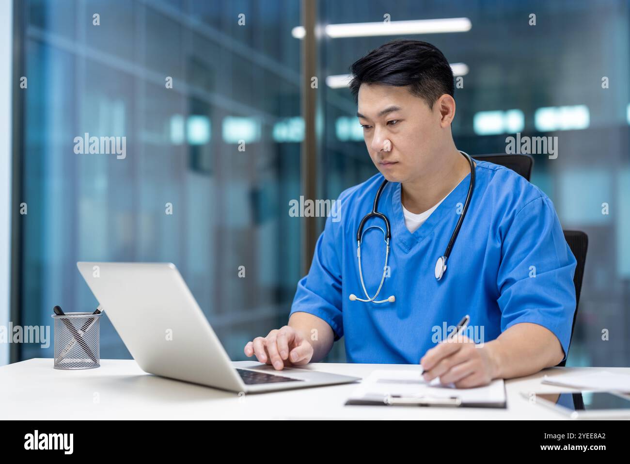 Seriöser asiatischer Arzt in Blau schrubbt mit Laptop und macht Notizen am Schreibtisch im modernen Krankenhaus. Medizinischer Fachmann, der sich auf die Arbeit konzentriert, Engagement und Gesundheitskompetenz unter Beweis stellt. Stockfoto