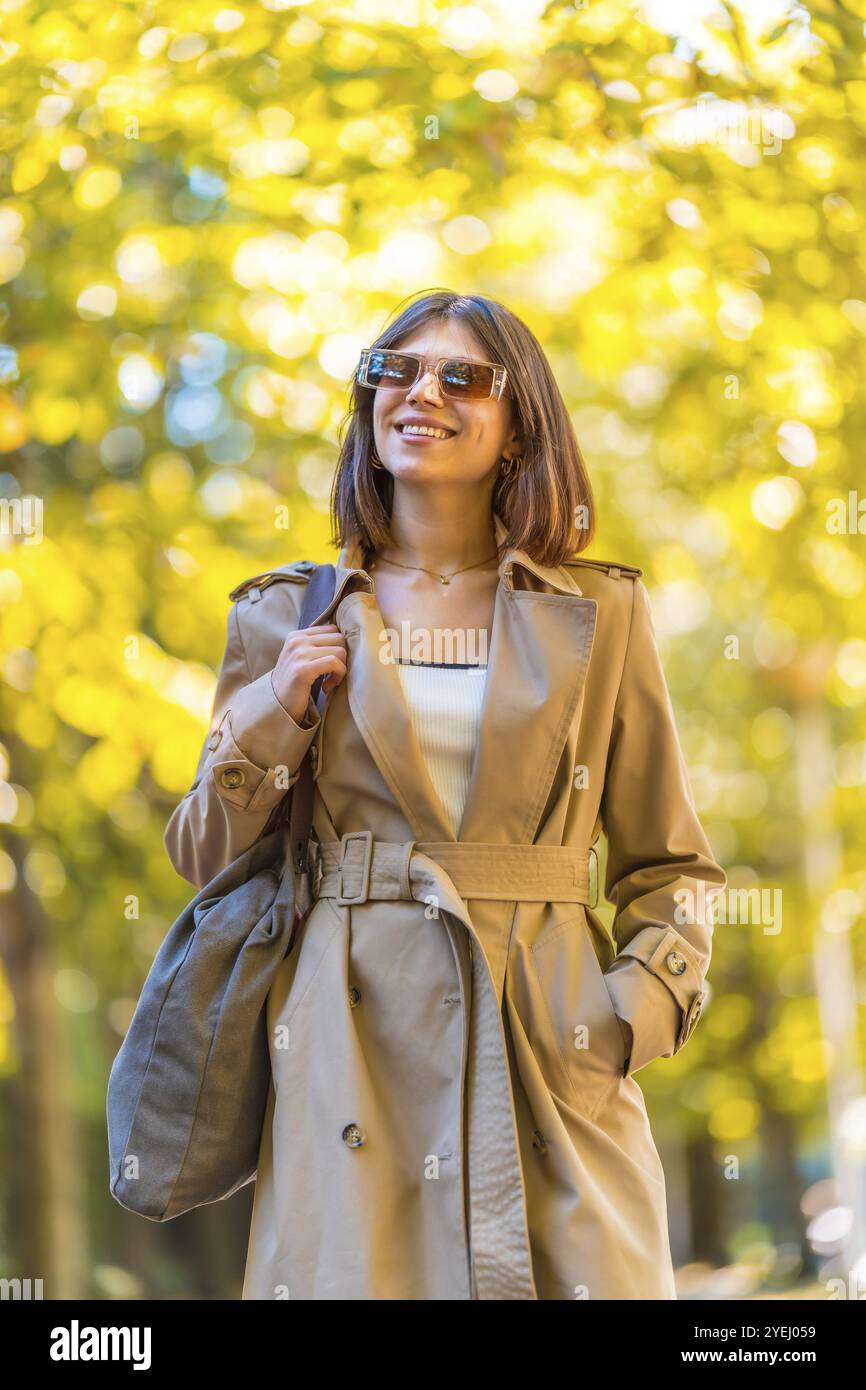 Eine Frau, die einen braunen Trenchcoat und eine Sonnenbrille trägt, läuft einen Weg entlang. Sie lächelt und hält eine Handtasche. Herbst- oder Winterkonzept in der Stadt Stockfoto