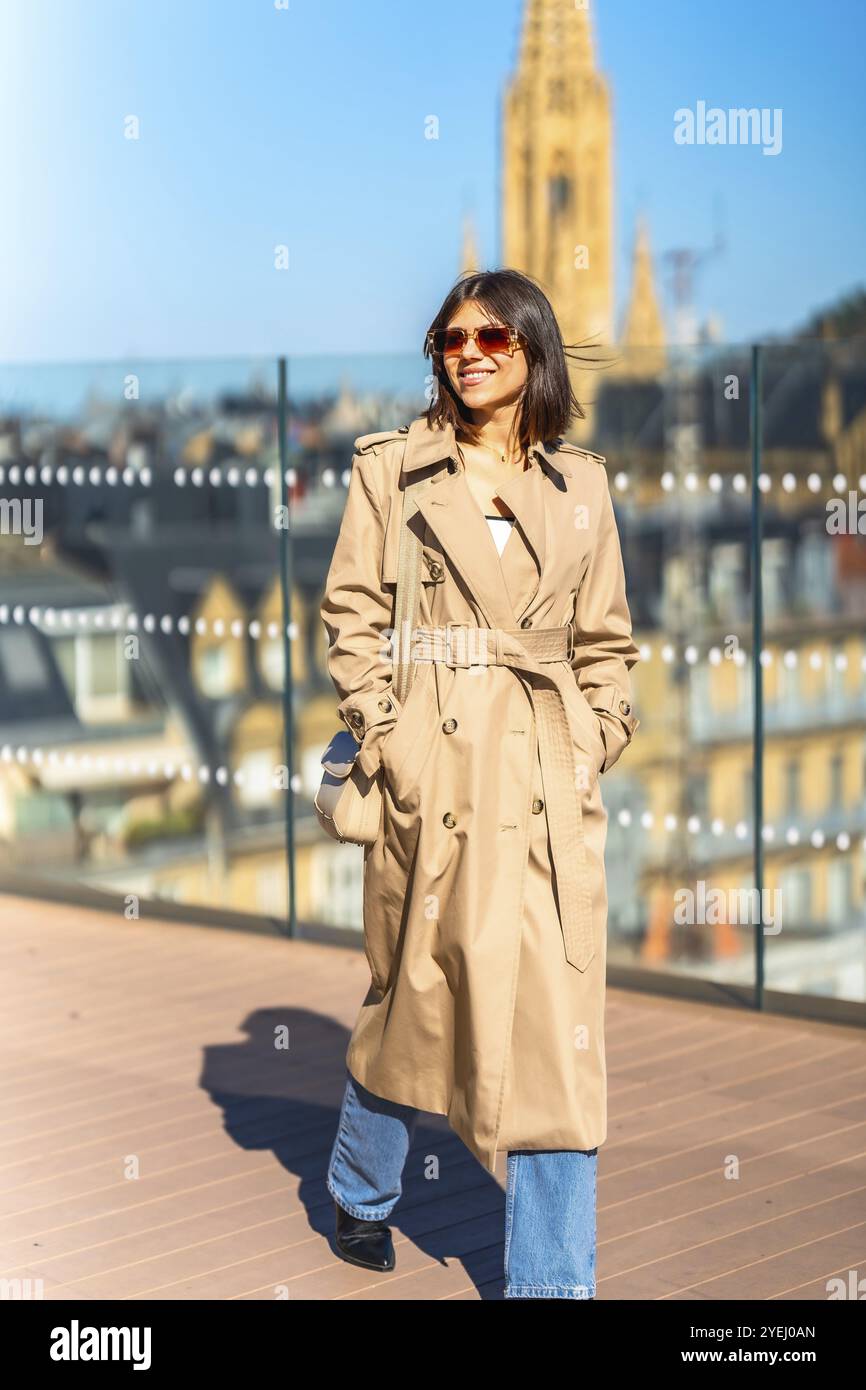 Eine Frau, die einen braunen Trenchcoat trägt, läuft auf einem Holzdeck. Sie trägt eine Handtasche und eine Sonnenbrille. Die Szene spielt in einer Stadt mit einem Tal Stockfoto