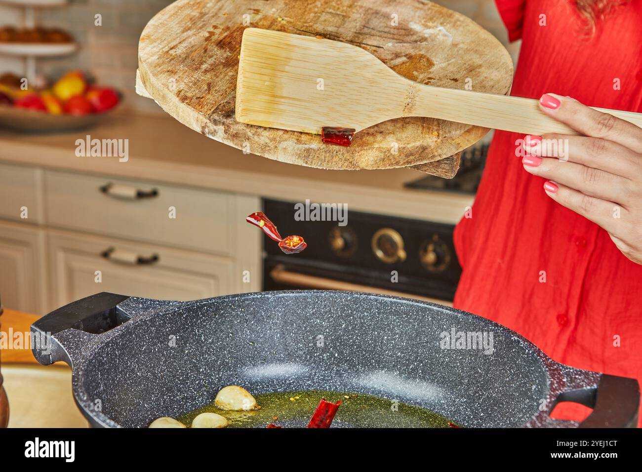 Eine Hand wirft getrocknete Chilistücke von einem Schneidebrett in eine Pfanne mit Knoblauch und Öl, um sie zum Kochen vorzubereiten. Stockfoto