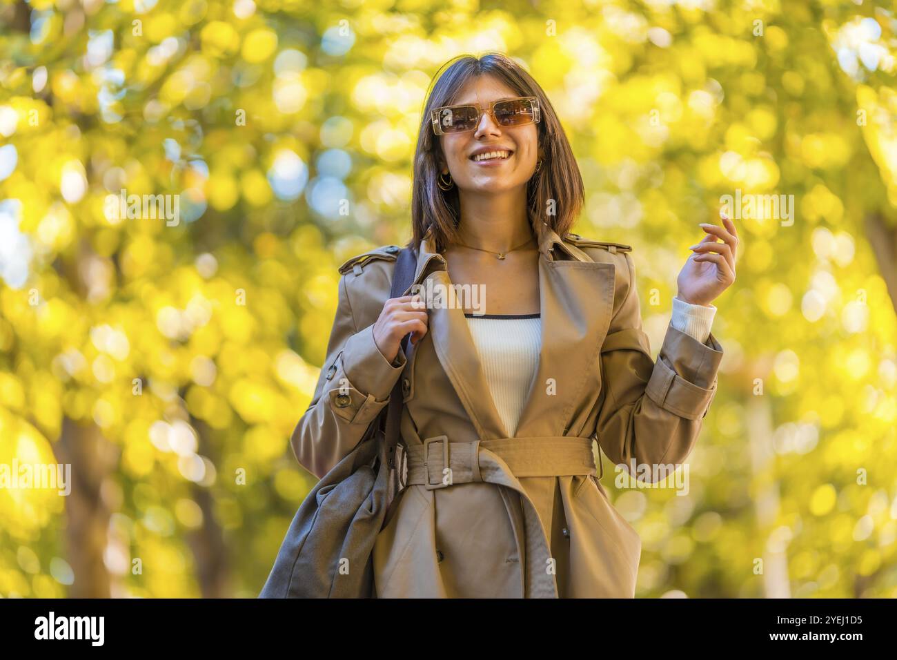 Eine Frau, die einen braunen Mantel und eine Sonnenbrille trägt, läuft einen Weg entlang. Sie lächelt und hält eine Handtasche. Herbst- oder Winterkonzept in der Stadt Stockfoto