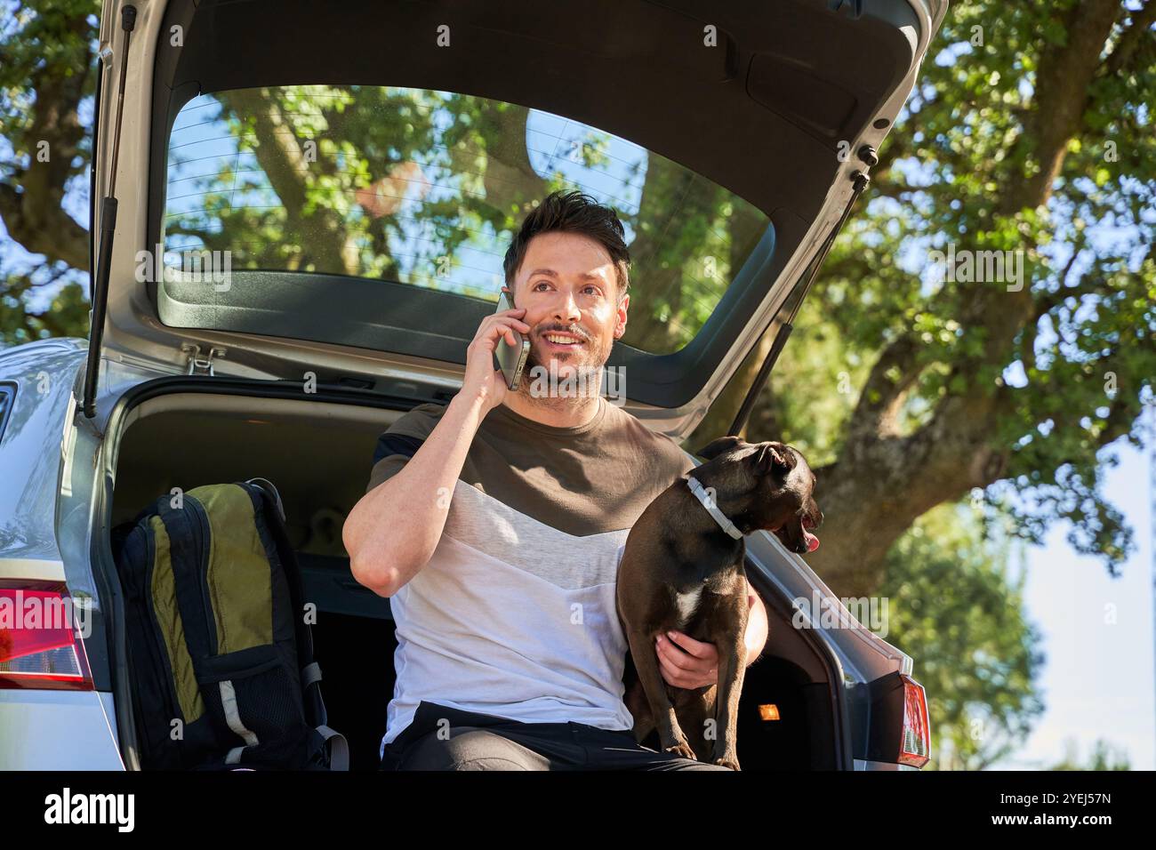 Mann sitzt im Kofferraum und spricht am Telefon. Er hat einen dunklen Hund in den Armen. Stockfoto