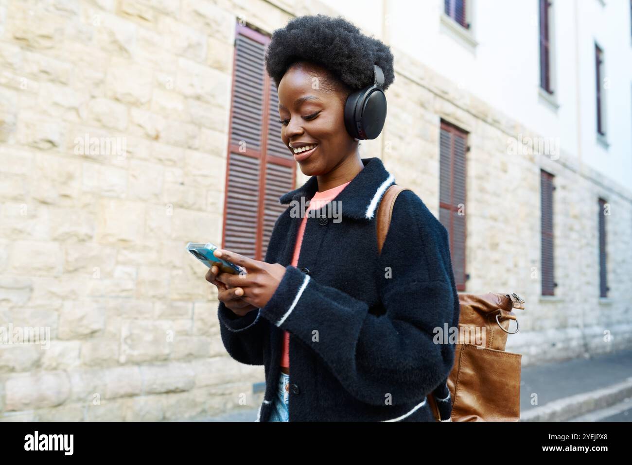 Junge Frau, die Musik über Kopfhörer genießt, während sie in der Stadt spaziert und ihr Smartphone überprüft Stockfoto