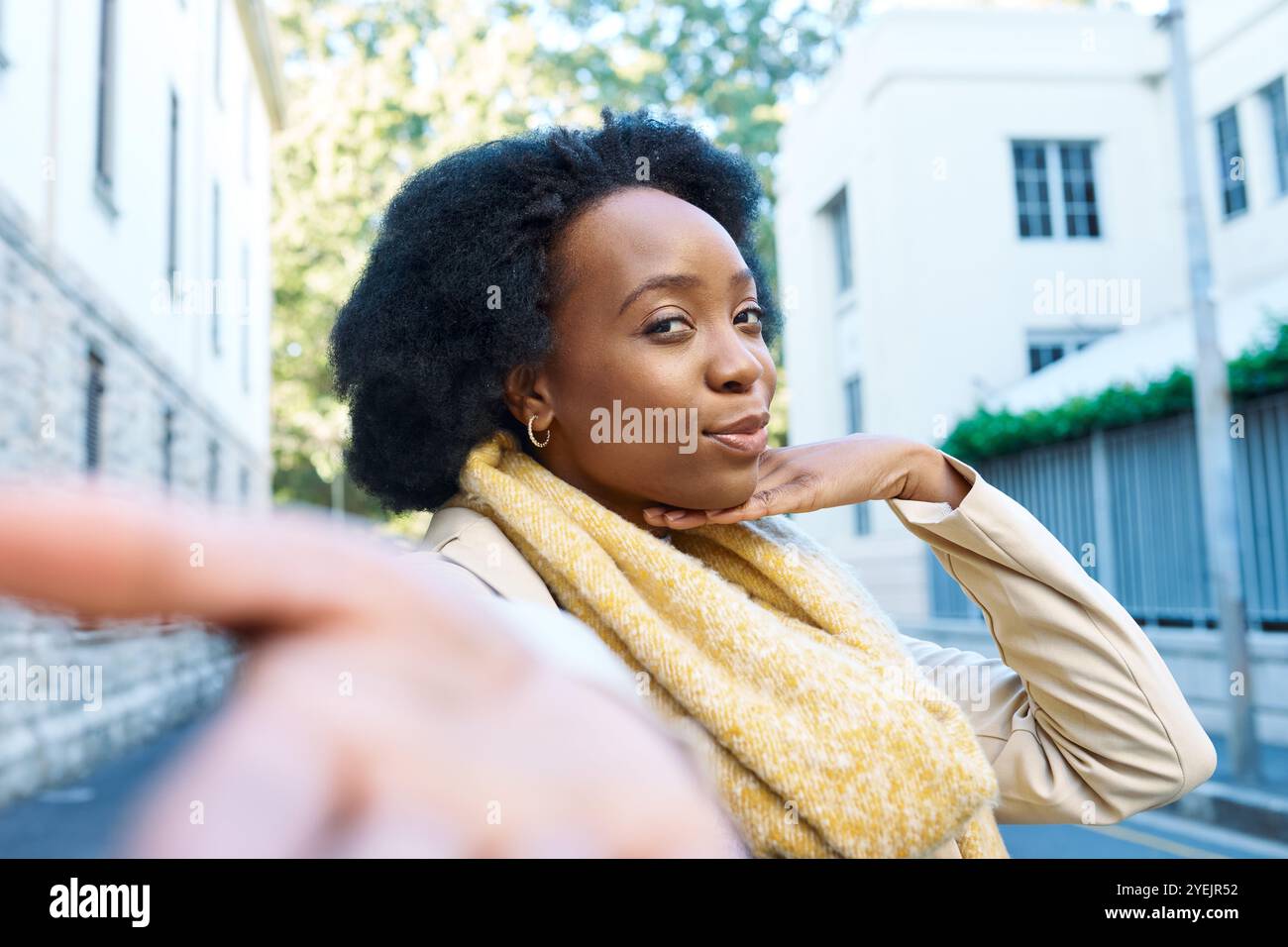 Selbstbewusste Frau, die an einem sonnigen Tag in der Stadt lächelt, trägt einen stylischen gelben Schal und eine beige Jacke und legt legere urbane Mode an Stockfoto