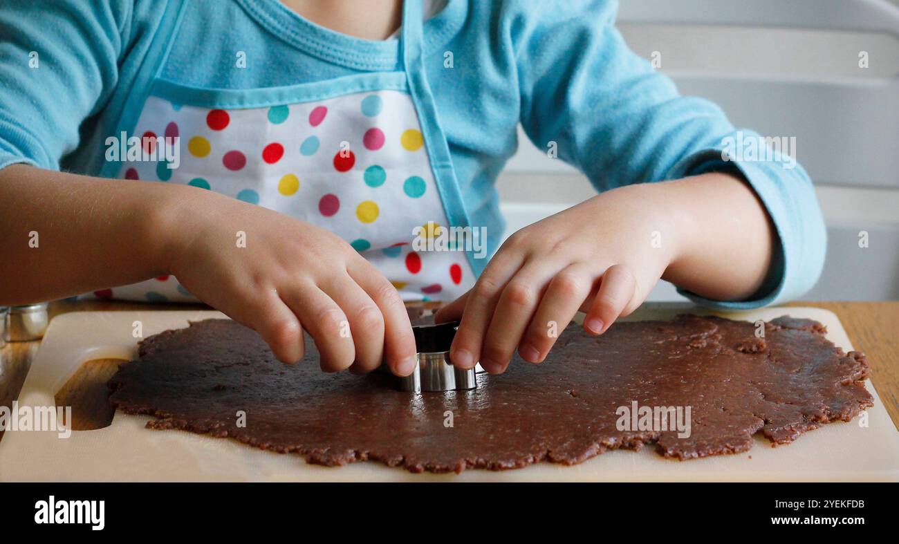 Hände eines 4-jährigen Kaukasiers in einer Schürze, die aus gerolltem Lebkuchenteig verschiedene Formen mit Weihnachtsmetallschneidern schneidet. Der Prozess von Stockfoto